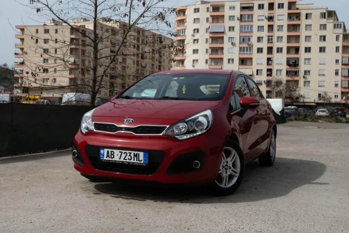 Front three-quarter view of red Kia Rio 2014 1.2 petrol manual rental car parked in front of apartment buildings in Tirana