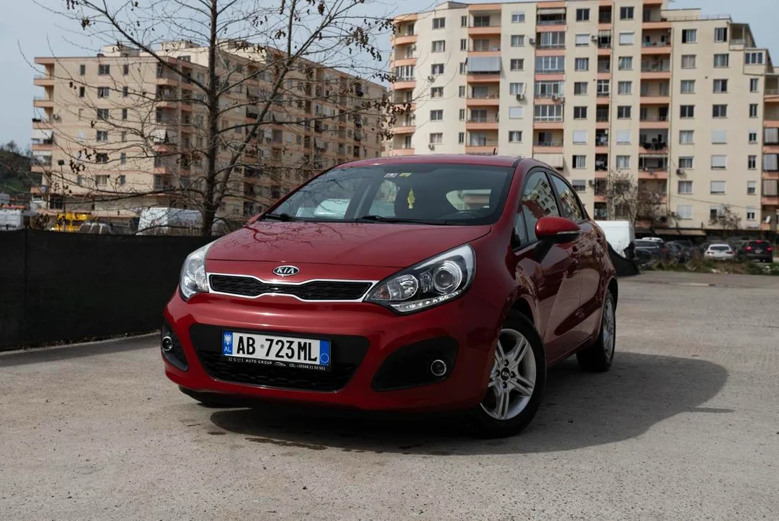 Kia Rio 2014 Rental in Tirana – 1.2 Petrol Manual City Car 36 - Car Rent in Albania Front three-quarter view of red Kia Rio 2014 1.2 petrol manual rental car parked in front of apartment buildings in Tirana